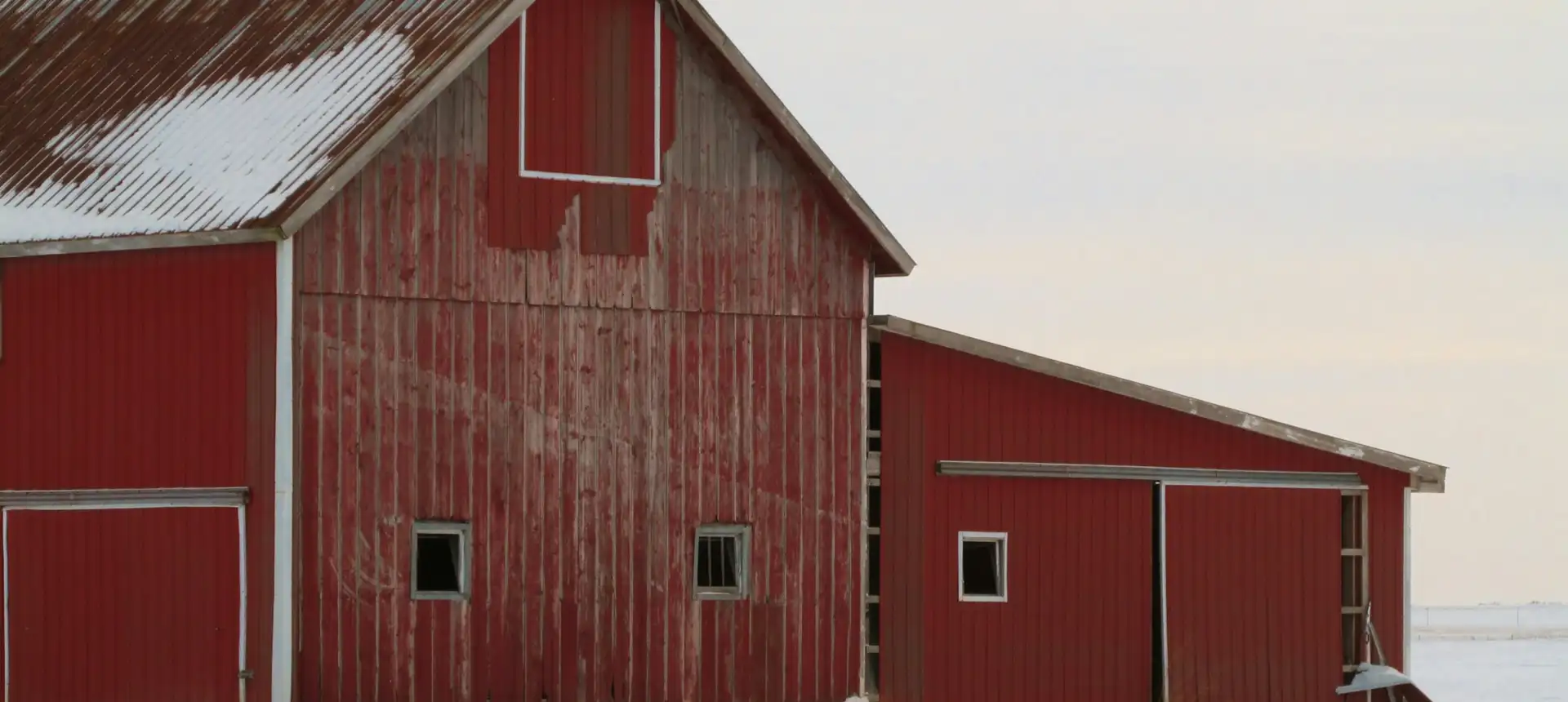 A red barn and the machine shed built onto it, standing in snow under a pale winter sky — the kind of farm outbuilding radiant floor heating is planned for before a slab is poured